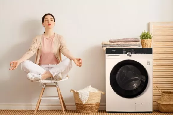 Vrouw zit te mediteren naast wasmachine omdat ze zich ontspannen voelt door een goede wasroutine